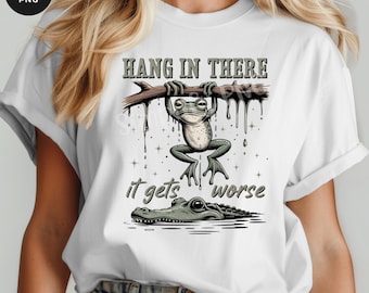 May include: White t-shirt with a cartoon frog clinging to a branch, the text "Hang in There" above and "it gets worse" below. The design includes a crocodile in the water.