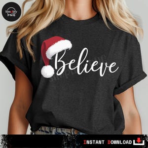Pode incluir: T-shirt cinzenta escura com a palavra "Believe" em escrita branca, acompanhada por um gráfico de gorro de Pai Natal. O gorro é vermelho com uma borda branca fofa e um pompom branco. Estilo de t-shirt casual.