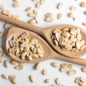 May include: Two wooden spoons filled with peeled peanuts on a white surface. The peanuts are scattered around the spoons.