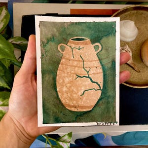 May include: Watercolor painting of a tan vase with a green crack design, set against a green background. The vase has two handles and is on a white paper. The painting is held in a hand.