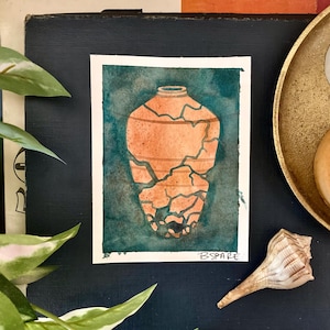 Può includere: Acquerello di un vaso color terracotta con crepe, su sfondo turchese. L'opera è su carta con bordi bianchi, firmata "BSPARE". Il dipinto è esposto con oggetti decorativi, tra cui una conchiglia.
