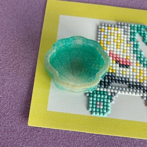 May include: A translucent, glittery, light green resin shell-shaped dish resting on a rectangular canvas featuring a partial diamond painting design with yellow, pink, teal, and grey square beads arranged in a grid pattern on a white background.