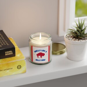 May include: A white candle in a glass jar with a red, white, and blue label that says "Smells Like A Bills Win!" with a red buffalo silhouette. The candle is lit and the flame is visible.