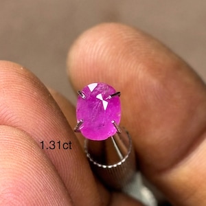 May include: A vibrant, oval-cut, pink gemstone, held in a silver setting. The gem is a deep magenta color, with visible internal structures. The text "1.31ct" is visible near the gem, indicating its weight in carats. The gem is held between fingers.