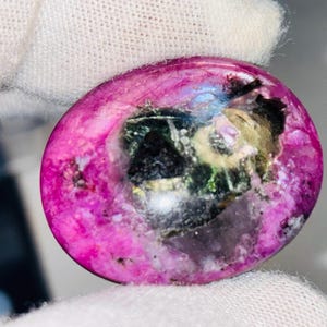 May include: A polished, oval-shaped gemstone with a vibrant magenta hue. The stone features a mix of colors, including black, green, and hints of yellow, creating a unique and eye-catching appearance. The gemstone is held by a gloved hand.