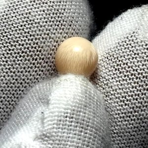 Può includere: Primo piano di un piccolo oggetto rotondo e chiaro, forse una perla, appoggiato su un tessuto bianco e strutturato. L'oggetto ha una superficie liscia e riflettente.