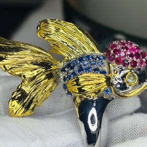 Op de afbeelding: Een goudkleurige ring in de vorm van een vis met ingewikkelde details. De vis heeft gouden vinnen, een kop versierd met rode edelstenen en een lichaam versierd met blauwe edelstenen. De ring is gezet op een zilveren band.