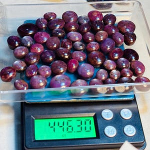 Puede incluir: Un recipiente de plástico transparente lleno de numerosas piedras preciosas pulidas, redondas y de color púrpura oscuro. El recipiente descansa sobre una balanza digital negra con una pantalla verde que muestra "446.30 ct". Se ven piedras preciosas adicionales.