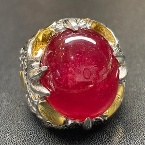 Può includere: Un anello ornato con una grande gemma rossa rotonda e brillante. La montatura combina metalli argentati e dorati, con dettagli intricati attorno alla gemma. Il design è audace e accattivante.