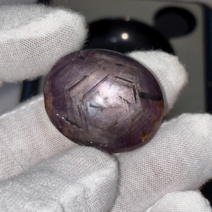 Peut inclure: Une pierre précieuse polie de forme ovale et de couleur violet foncé. La pierre présente un motif géométrique gravé à sa surface, avec des lignes et des facettes visibles. Le bijou est tenu dans une main gantée de blanc.