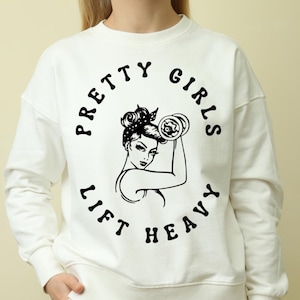 May include: White sweatshirt with a black graphic. The graphic features a woman lifting a dumbbell, surrounded by the text "PRETTY GIRLS LIFT HEAVY" in a circular design. The woman has a polka dot headscarf.