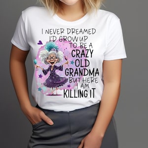 以下が含まれることがあります： ワイングラスを持ったおばあちゃんの漫画イラストが描かれた白いTシャツ。「I NEVER DREAMED I'D GROW UP TO BE A CRAZY OLD GRANDMA BUT HERE I AM KILLING IT.」と書かれています。