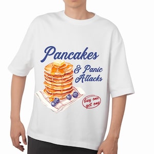 Könnte beinhalten: Weißes T-Shirt mit dem blauen Schriftzug "Pancakes & Panic Attacks". Die Grafik zeigt einen Stapel Pfannkuchen mit Butter und Sirup, Blaubeeren und einen Kreis "buy one get one".