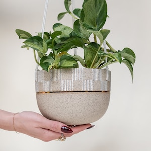 May include: A hanging planter with a beige and white geometric pattern and a gold trim. The planter is filled with a green and white variegated plant.