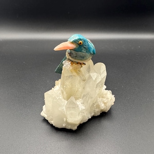 Op de afbeelding: Een decoratieve vogelsculptuur met een blauwe kop en roze snavel, geplaatst op een cluster van heldere kwartskristallen. De vogel heeft gele ogen en een gedetailleerd, gestructureerd uiterlijk. De kwartskristallen zijn samengeklonterd en vormen een natuurlijke, organische basis.