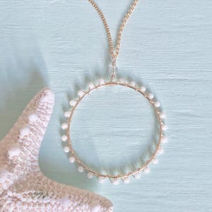 Op de afbeelding: Een gouden ketting met een witte ronde hanger gemaakt van kleine witte kralen. De hanger is met draad gewikkeld en heeft een delicate, luchtige uitstraling.