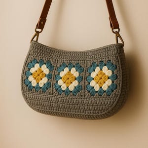 May include: A gray crocheted handbag with a brown leather strap. The bag features three floral designs in blue, yellow, and white. The bag has a curved shape and is suspended against a neutral background.