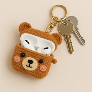 May include: A brown crocheted bear-shaped case for AirPods with a gold keyring attached. The case is open, revealing the white AirPods inside.