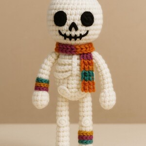 Könnte beinhalten: Weiße gehäkelte Skelettpuppe mit einem bunten gestreiften Schal und passenden Armbändern. Die Puppe hat schwarze Augen und einen lächelnden Mund.