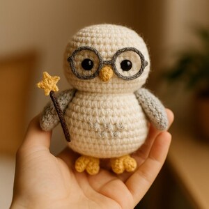 Peut inclure: Un hibou en crochet blanc portant des lunettes grises et tenant une baguette en bois avec une étoile jaune au sommet. Le hibou a des pieds jaunes et est tenu dans la main d'une personne.