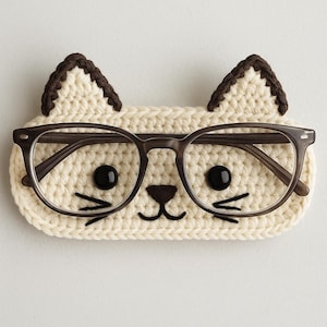 May include: A crocheted cat-shaped glasses holder with brown ears and a white face. The holder has black eyes, a brown nose, and white whiskers.