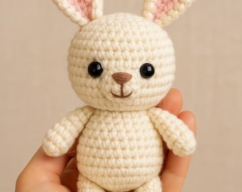 May include: A crocheted white bunny with pink ears and black eyes. The bunny is sitting on a hand.