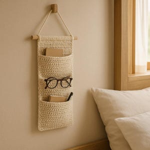 以下が含まれることがあります： 3つのポケットが付いたベージュのクロシェの壁掛けオーガナイザー。オーガナイザーは壁に掛けられており、上の2つのポケットには眼鏡とペンが入っています。