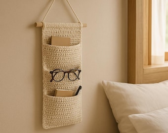 May include: A beige crocheted wall organizer with three pockets. The organizer is hanging on a wall and has a pair of glasses and a pen in the top two pockets.