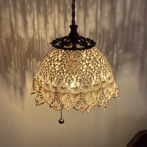 May include: A pendant light fixture featuring a beige, intricate crochet lace shade with a scalloped edge. The shade is attached to a dark brown, ornate metal cap and a twisted cord, with a small pull chain hanging from the center of the light.