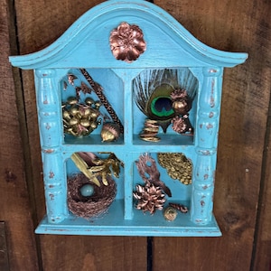 May include: A turquoise-painted, vintage-style shadow box with four compartments. The box is adorned with copper-coloured embellishments and filled with natural elements like a peacock feather, a bird's nest, and gilded acorns. The wooden background adds a rustic touch.