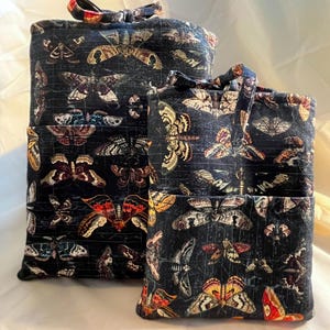 May include: Two rectangular fabric pouches in varying sizes, featuring a dark black background with a repeating pattern of colorful moths and butterflies in shades of orange, yellow, white, and brown. Each pouch has a top handle and a front pocket.