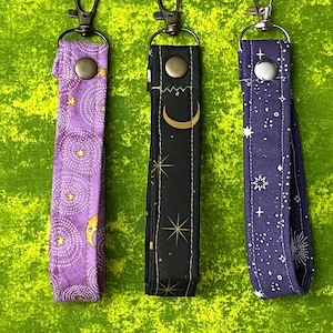 May include: Three fabric wristlet keychains with metal lobster clasps and snap buttons. The left is purple with yellow moons and stars, the center is black with gold celestial patterns, and the right is dark blue with white star constellations.