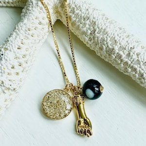 May include: Gold chain necklace with three charms: a round charm filled with sand, a gold hand charm, and a black and white agate bead.