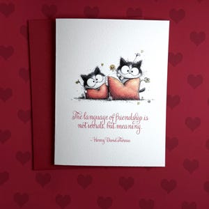 May include: A greeting card with a red envelope, featuring a whimsical illustration of two black and white cats on red pillows. The card includes the quote, "The language of friendship is not words, but meaning."