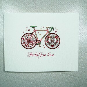 May include: A greeting card featuring a pink bicycle illustration with floral and arrow accents. The wheels are decorated with hearts and flowers. The text "Pedal for love" is written in a cursive font below the bicycle.