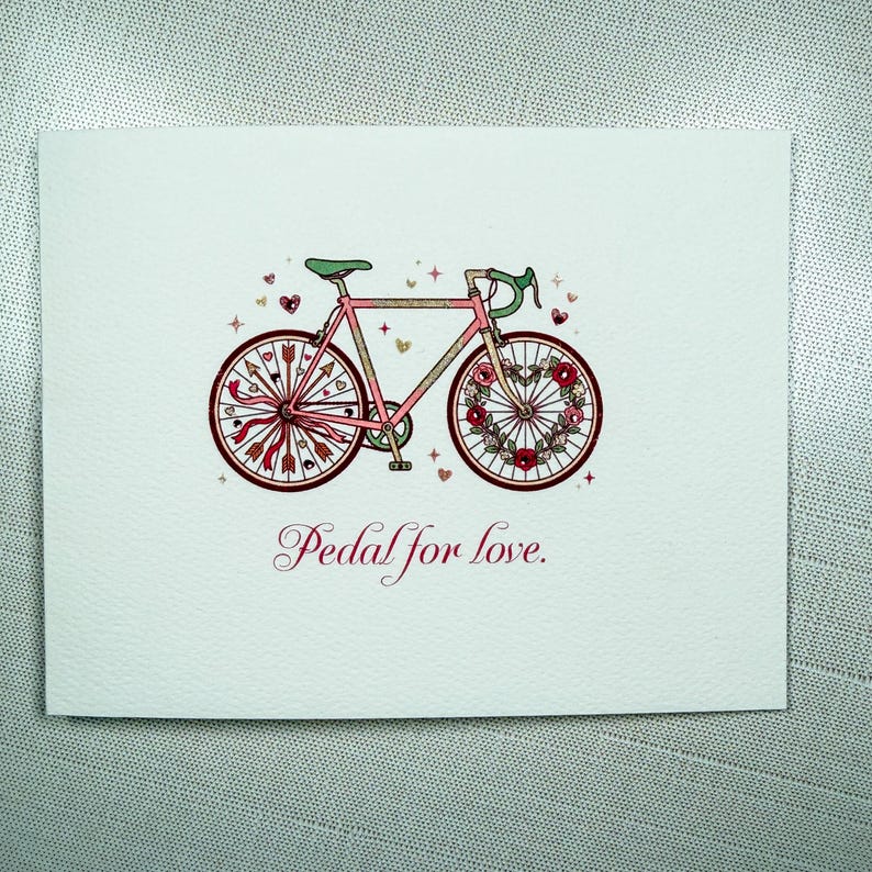May include: A greeting card featuring a pink bicycle illustration with floral and arrow accents. The wheels are decorated with hearts and flowers. The text "Pedal for love" is written in a cursive font below the bicycle.