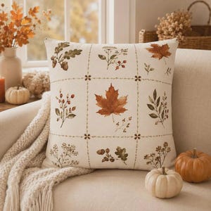 May include: A square throw pillow with an off-white base featuring a grid pattern. Each section contains botanical illustrations of autumn elements including maple leaves, oak leaves with acorns, berries, and dried wildflowers in shades of brown.
