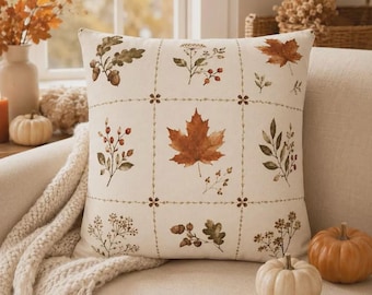 May include: A square throw pillow with an off-white base featuring a grid pattern. Each section contains botanical illustrations of autumn elements including maple leaves, oak leaves with acorns, berries, and dried wildflowers in shades of brown.