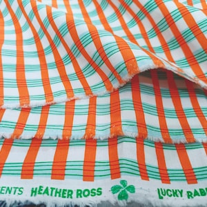 May include: A white fabric featuring a plaid pattern of bold orange vertical stripes and thinner green and black horizontal lines. The selvage edge displays the green printed text: PRESENTS HEATHER ROSS LUCKY RABBIT with a small clover icon.
