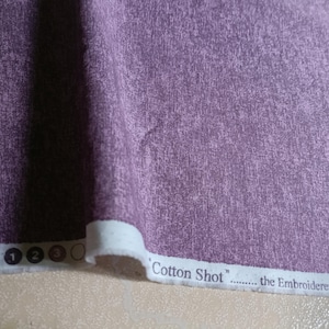 Może przedstawiać: Zbliżenie fioletowej tkaniny o teksturowanym, nakrapianym wyglądzie. Tkanina jest oznaczona jako "Cotton Shot" i "the Embroiderer's & Quilter's". Tkanina ma miękki, lekko wyblakły wygląd, odpowiedni do pikowania lub innych projektów tekstylnych.