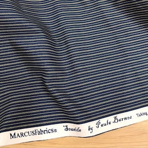 Könnte beinhalten: Nahaufnahme eines marineblauen Stoffes mit dünnen, horizontalen, cremefarbenen Streifen. Der Stoff ist leicht zerknittert, was darauf hindeutet, dass es sich um ein weiches Material handelt. Der Text "MARCUS Fabrics Seaside by Paula Barnes Ticking R225" ist am unteren Rand aufgedruckt.