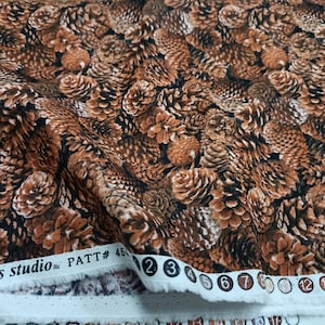 Puede incluir: Primer plano de tela con un patrón repetido de piñas marrones y negras. La tela está doblada, mostrando su textura. El borde de la tela muestra el texto "elizabeth's studio inc PATT# 45" y números.