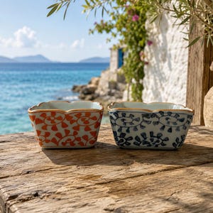 May include: Two small square ceramic bowls with scalloped edges. One bowl features an orange repeating floral pattern, while the other displays a blue repeating floral pattern. Both bowls have a white base color and a thin brown rim along the top edge.