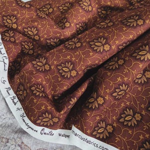 May include: A folded piece of fabric featuring a dark reddish-brown background with a repeating pattern of golden-yellow stylized flowers and thin, winding vines. The white selvage edge displays the text Pam Buda of Heartspun Quilts Wallpar marcusfabrics.com.