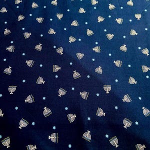 Peut inclure: Tissu bleu marine avec un motif répété de menorahs blanches et d'étoiles bleu clair. Les menorahs ont un contour doré et des détails verts. Le tissu est étiqueté 100% coton.