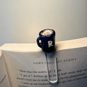 Op de afbeelding: Een blauwe keramische mok-bladwijzer met wit glazuur erop. De bladwijzer is gevormd als een mok met een handvat en een letter "R" aan de zijkant. De bladwijzer ligt op een open boek met de zichtbare tekst "NORA GOES OFF SCRIPT".