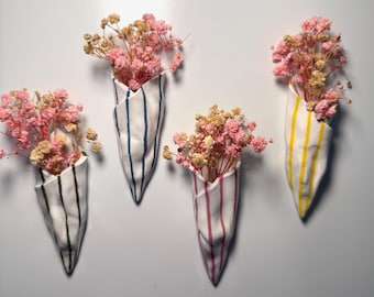 Op de afbeelding: Vier witte keramische wandvazen met verticale strepen in verschillende kleuren, elk met gedroogde roze en bruine bloemen. De vazen zijn kegelvormig en aan een witte muur bevestigd.