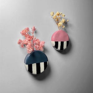 May include: Two ceramic wall vases, one navy blue and one pink, each with a black and white striped design on the bottom half. The vases hold dried flowers, adding a touch of natural beauty to the modern decor.
