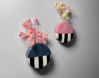 Op de afbeelding: Twee keramische wandvazen, één marineblauw en één roze, elk met een zwart-wit gestreept ontwerp op de onderste helft. De vazen bevatten gedroogde bloemen, wat een vleugje natuurlijke schoonheid toevoegt.