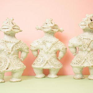 May include: Three off-white ceramic figurines with detailed facial features and ornate clothing. The figures stand with their arms at their sides, and have a unique, stylized appearance. The background is a soft pink and green.